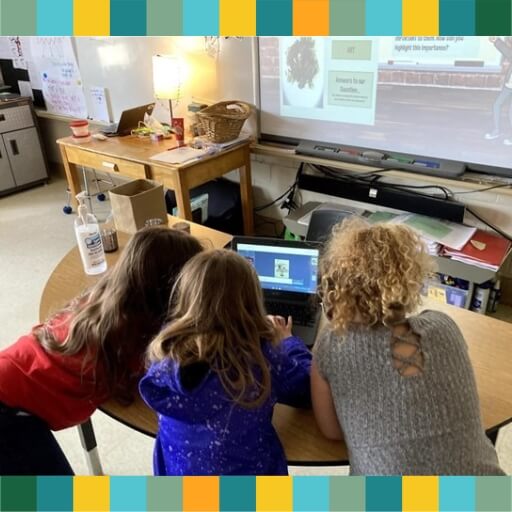 Three people gathered around a laptop in a classroom setting.